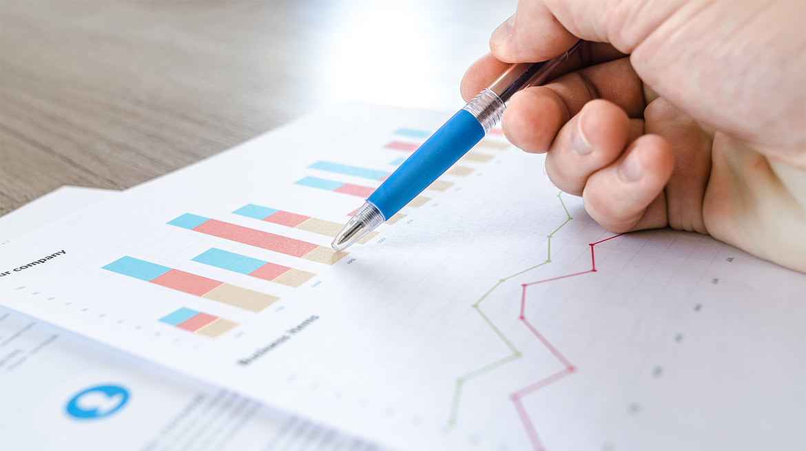 Hand mit Stift die über einem Blatt mit Statistikzahlen zeigt.